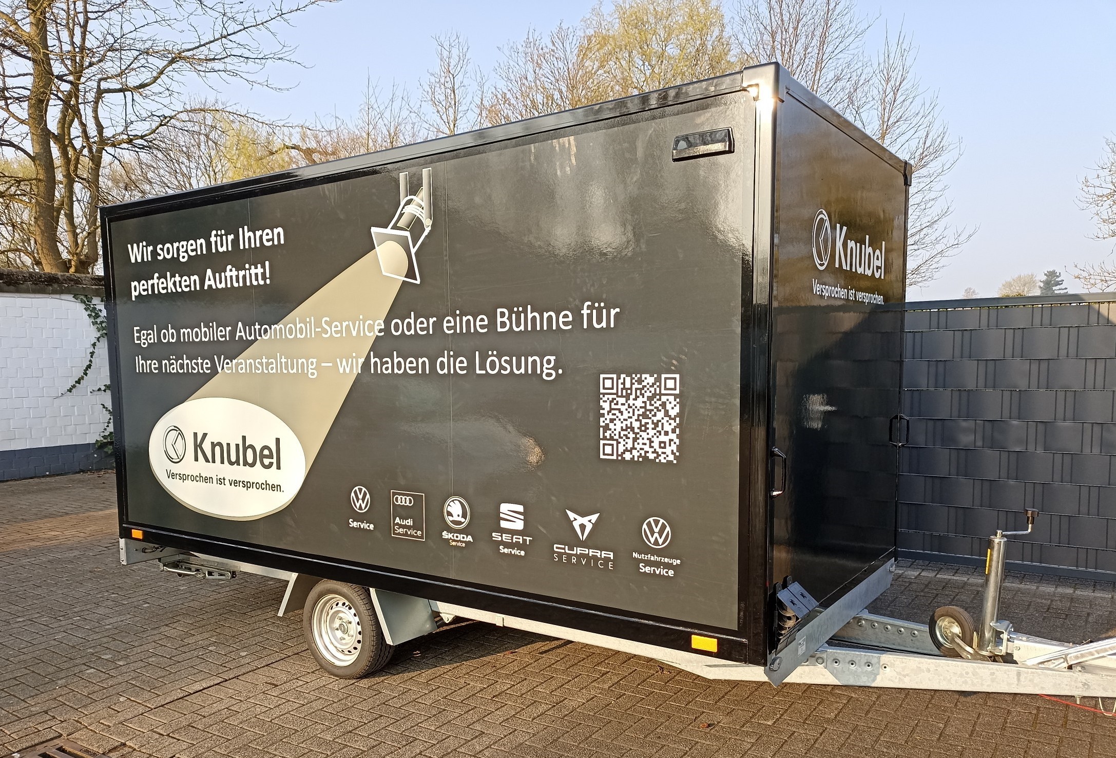 Der Anh&auml;nger f&uuml;r die B&uuml;hne des Knubel Service mobils mit der Aufschrift "Wir sorgen f&uuml;r Ihren Auftritt! Egal ob mobiler Automobil-Service oder eine B&uuml;hne f&uuml;r Ihre n&auml;chste Veranstaltung - wir haben die L&ouml;sung."